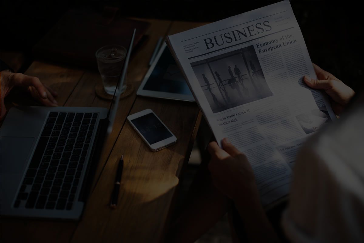 primeiro slide - Sobre uma mesa de madeira, uma pasta em couro, um notebook, um copo de vidro com água e gelo, um tablet, um telefone e uma caneta. Em frente à mesa e sentada uma mulher branca segura um jornal com a manchete Business